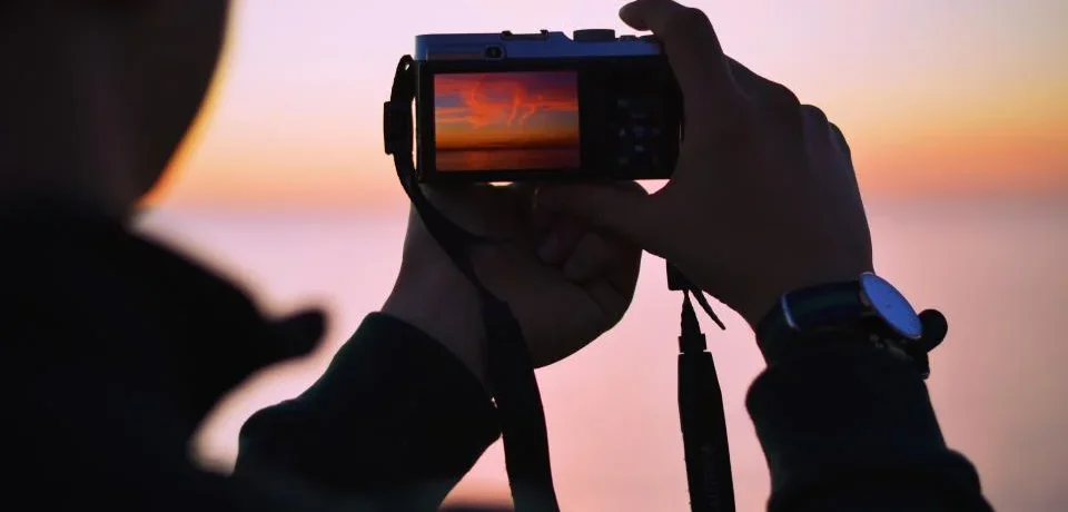Person photographing a clean car with a smartphone during golden hour against a plain background