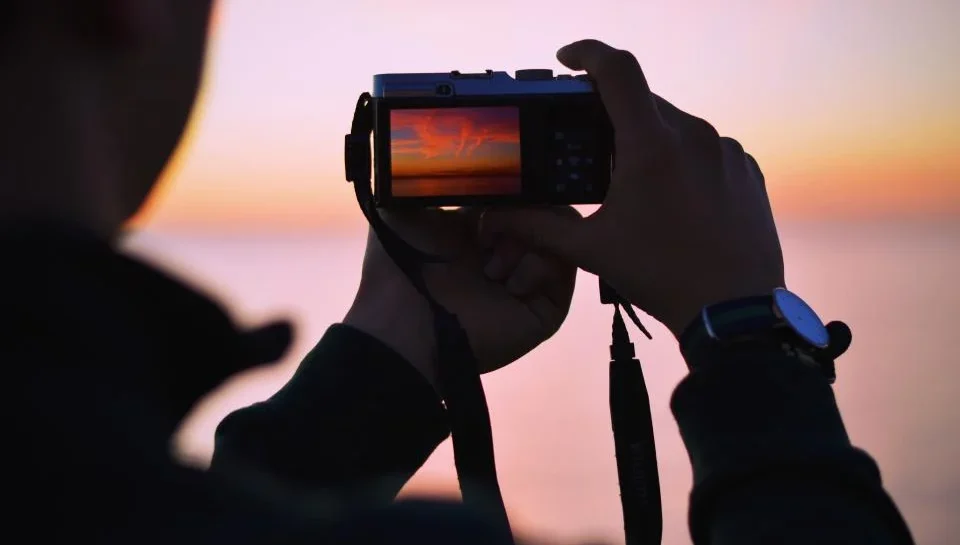 Person photographing a clean car with a smartphone during golden hour against a plain background